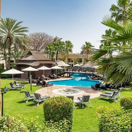 Le Meridien Dubai Hotel & Conference Centre Exterior foto The photo shows a serene outdoor scene featuring a swimming pool surrounded by lush greenery. There are several palm trees and shrubs providing a tropical atmosphere. Lounge chairs are arranged around the pool, along with shaded areas created by umbr