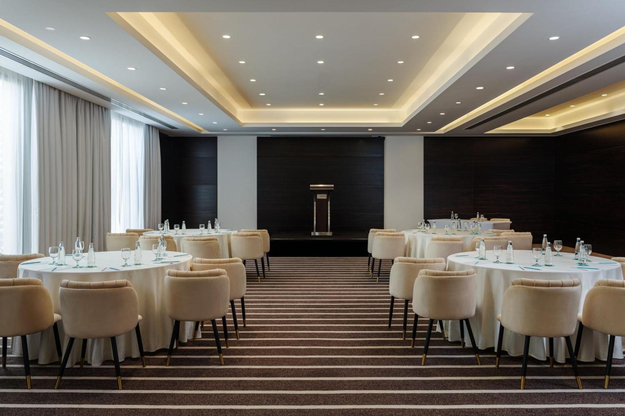 Le Meridien Dubai Hotel & Conference Centre Exterior foto The photo shows an elegantly designed conference or banquet room. There are round tables covered with white tablecloths, each accompanied by plush, light-colored chairs. The flooring features a striped pattern. The room is well-lit with recessed ceil
